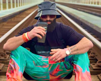 Nevv Evve sitting on railroad tracks and holding tea while wearing colorful and artistic luxury streetwear with designer accessories and a Loewe bucket hat during an early morning songwriting session.