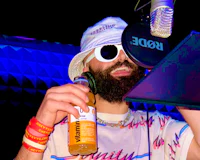 Nevv Evve recording vocals in a professional music studio during a custom song creation session while wearing artistic clothing with white sunglasses and holding a Vitamin Water in a neon-lit creative environment.